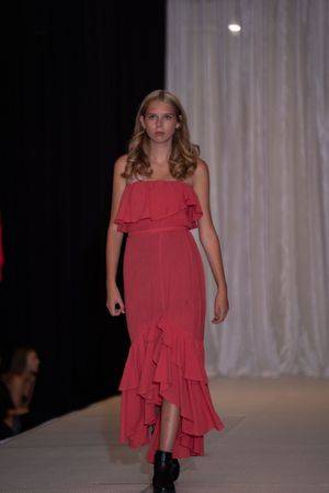 Model walks runway wearing flowing coral dress with ruffled layers during fashion show.