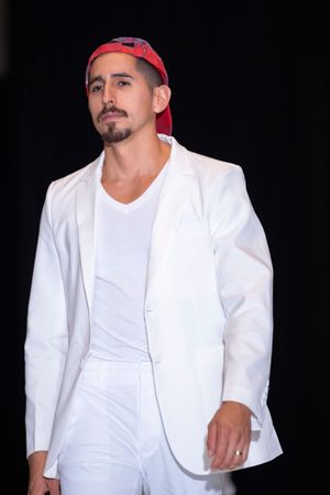 Model in all-white suit and red baseball cap poses on dark runway.