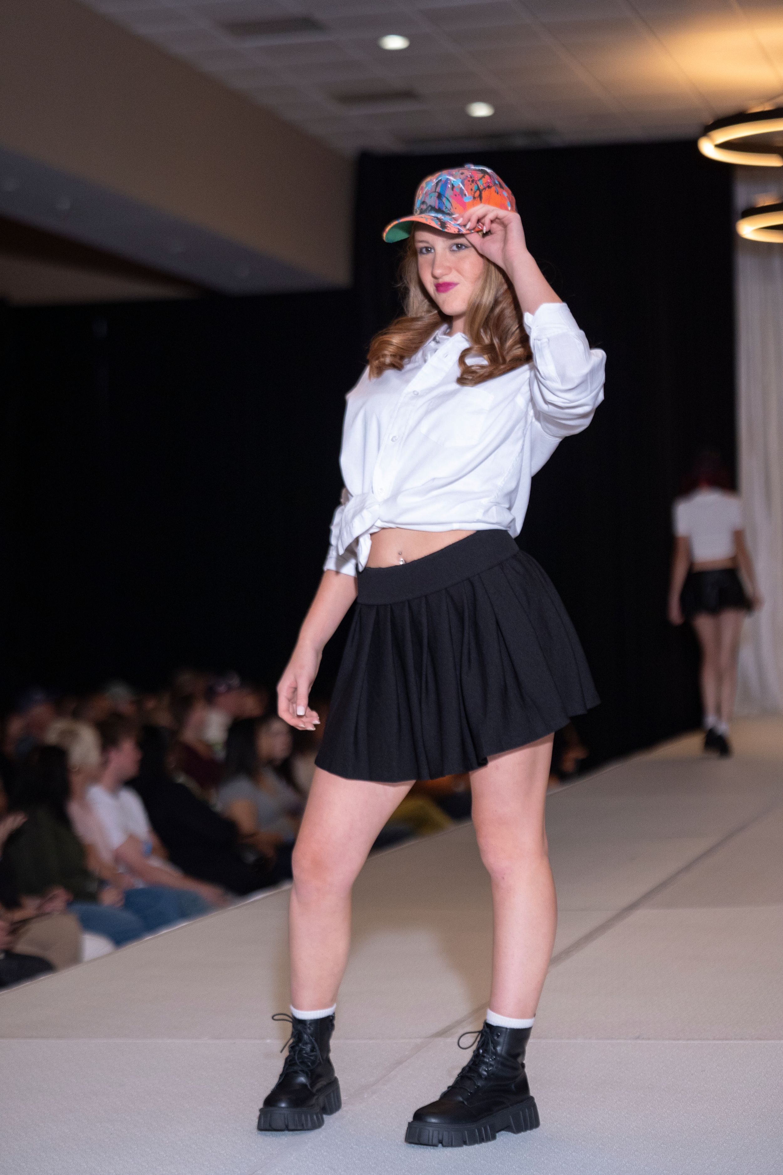 Model poses in black skirt and white top with combat boots on catwalk.