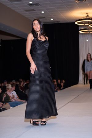 Model walks runway in black striped evening dress during indoor fashion show presentation.