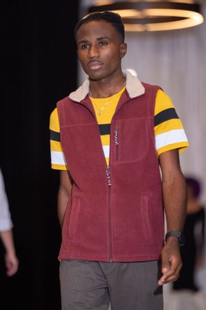 Model wears burgundy fleece vest with yellow striped t-shirt on fashion runway.