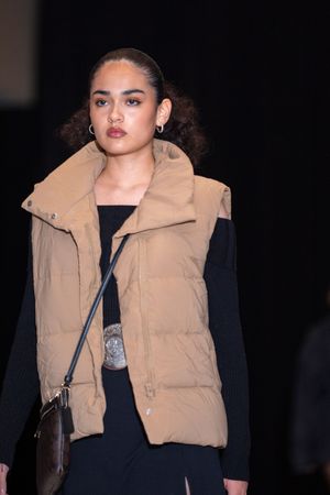 Model showcases tan puffer vest with black sweater and crossbody bag on runway.