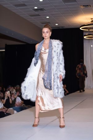 Model walks runway in white fur coat and neutral dress during fashion show.