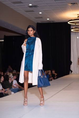 Model presents blue sleeveless dress with white jacket on fashion runway.