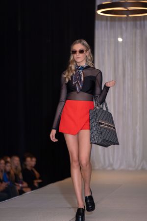 Model presents sheer black top with red shorts and quilted handbag on catwalk.