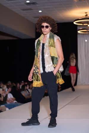 Model walks runway in black outfit with gold and white scarf accessories at fashion show.