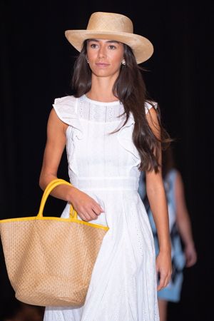 Fashion model walks runway in white summer dress with straw hat and yellow tote bag at fashion show.