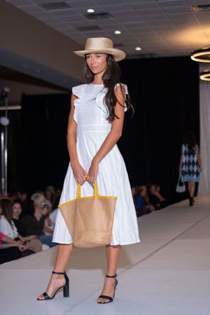 Fashion model walks runway in white summer dress and tan handbag at indoor fashion show.