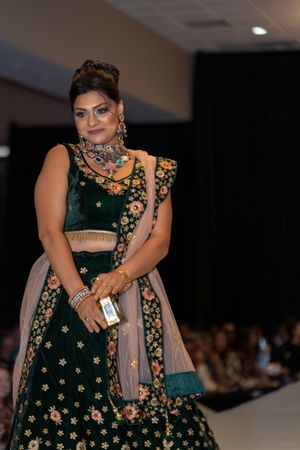Model walks runway in emerald green lehenga with gold embroidery and beige dupatta at fashion show.