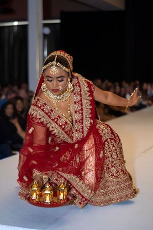 Model showcases ornate red and gold traditional Indian bridal lehenga on fashion runway with decorative candles.