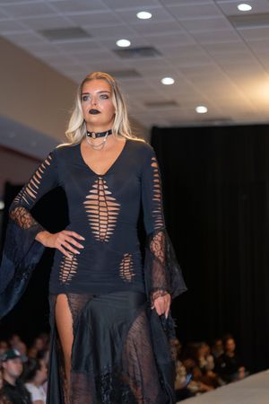 Model walks runway in sheer black lace evening gown with long sleeves during fashion show.