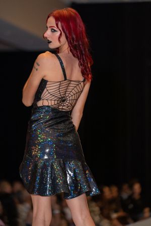 Model walks runway in sparkly holographic mini dress with spider web-inspired back detail at fashion show.
