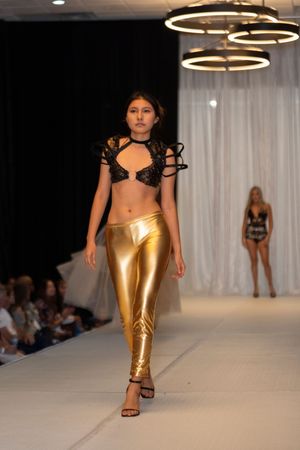 Fashion model walks runway in metallic gold pants and black top at fashion show event.