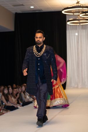Model showcases navy blue traditional outfit with gold accessories on illuminated runway.