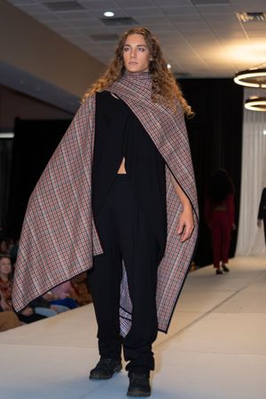 Model walks runway wearing black ensemble with plaid cape overlay during fashion show.