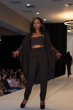 Model in all-black athleisure outfit strikes dynamic pose on white runway during fashion show.
