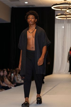 Model walks runway wearing black open shirt and pants with loafers during fashion show.