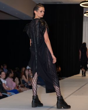Model showcases black dress with fishnet stockings on fashion runway.