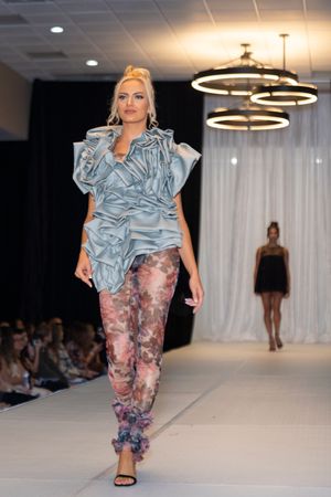 Model showcases light blue ruffled top with floral print pants on fashion runway.