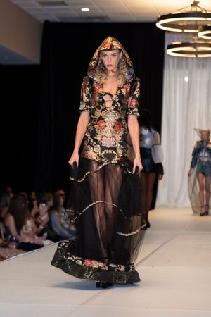 Model showcases dramatic hooded floral gown with sheer black overlay on fashion runway.