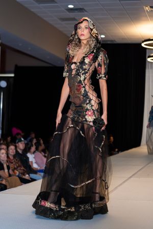Model walks in sheer black floral gown with dramatic details on catwalk.