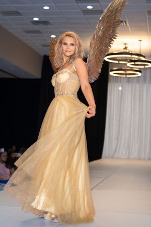 Model wearing gold evening gown with large feathered wings walks fashion runway.