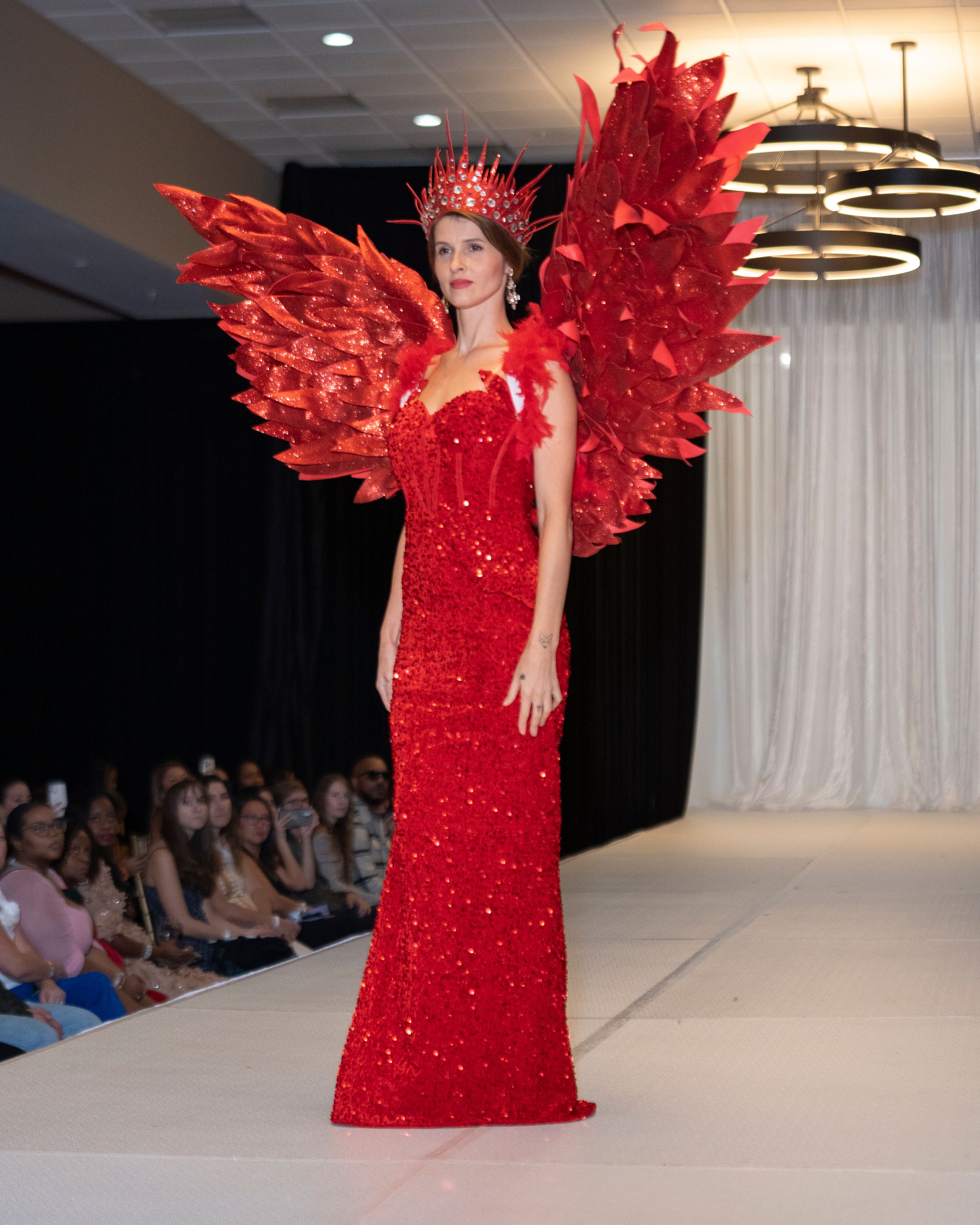 Dramatic red sequined gown with feathered wings displayed on fashion runway.