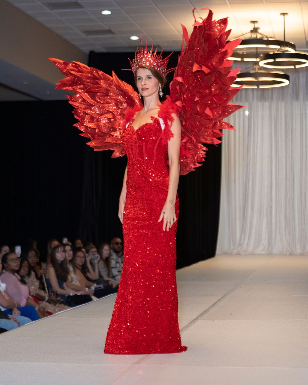 Dramatic red sequined gown with feathered wings displayed on fashion runway.