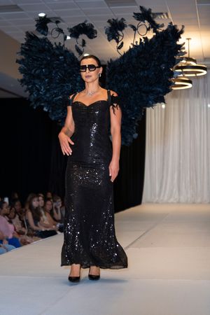 Model presents sparkly black evening gown with dramatic feathered shoulders on runway.