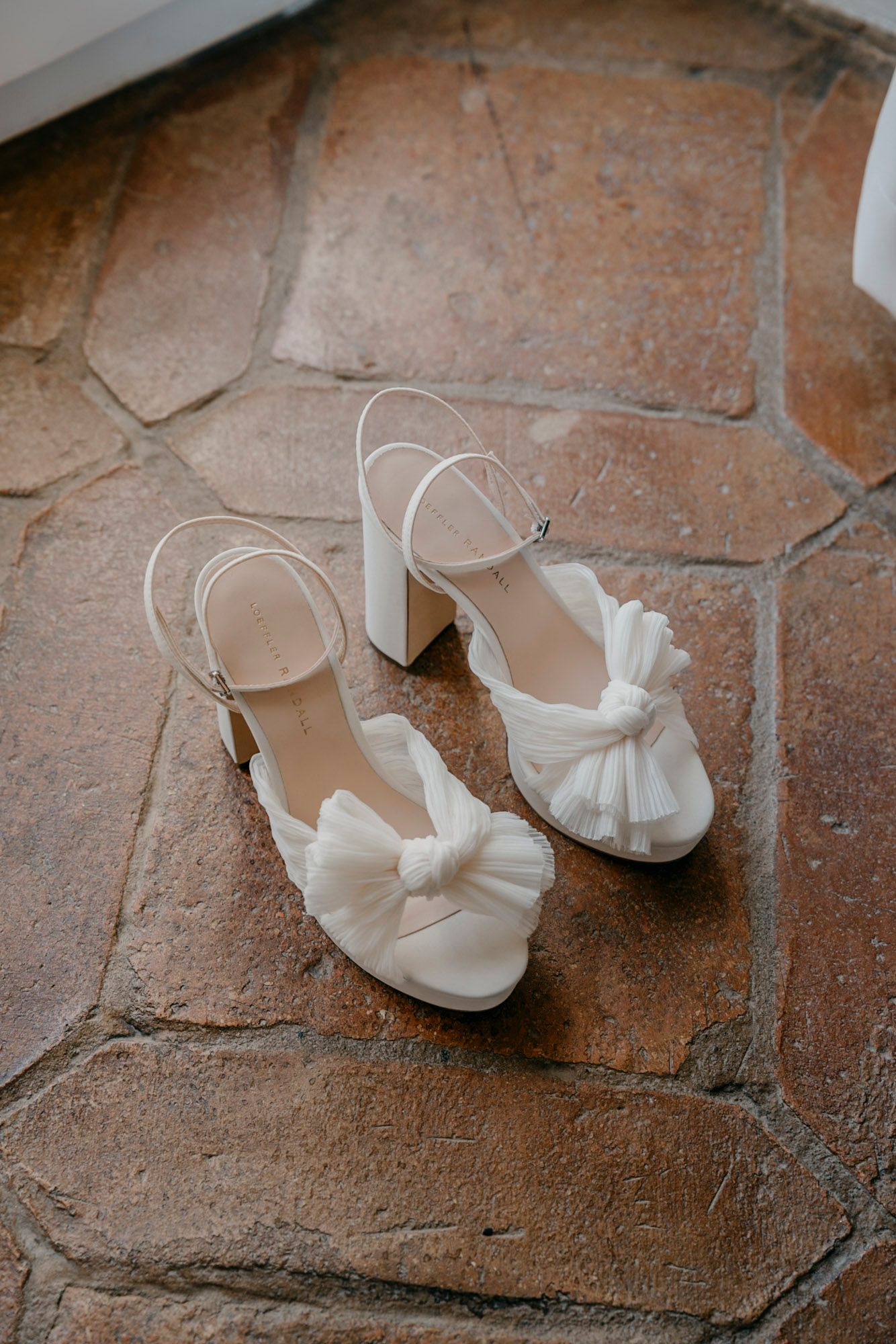 Elegant white bridal shoes with bow details and block heels placed on terracotta floor tiles.