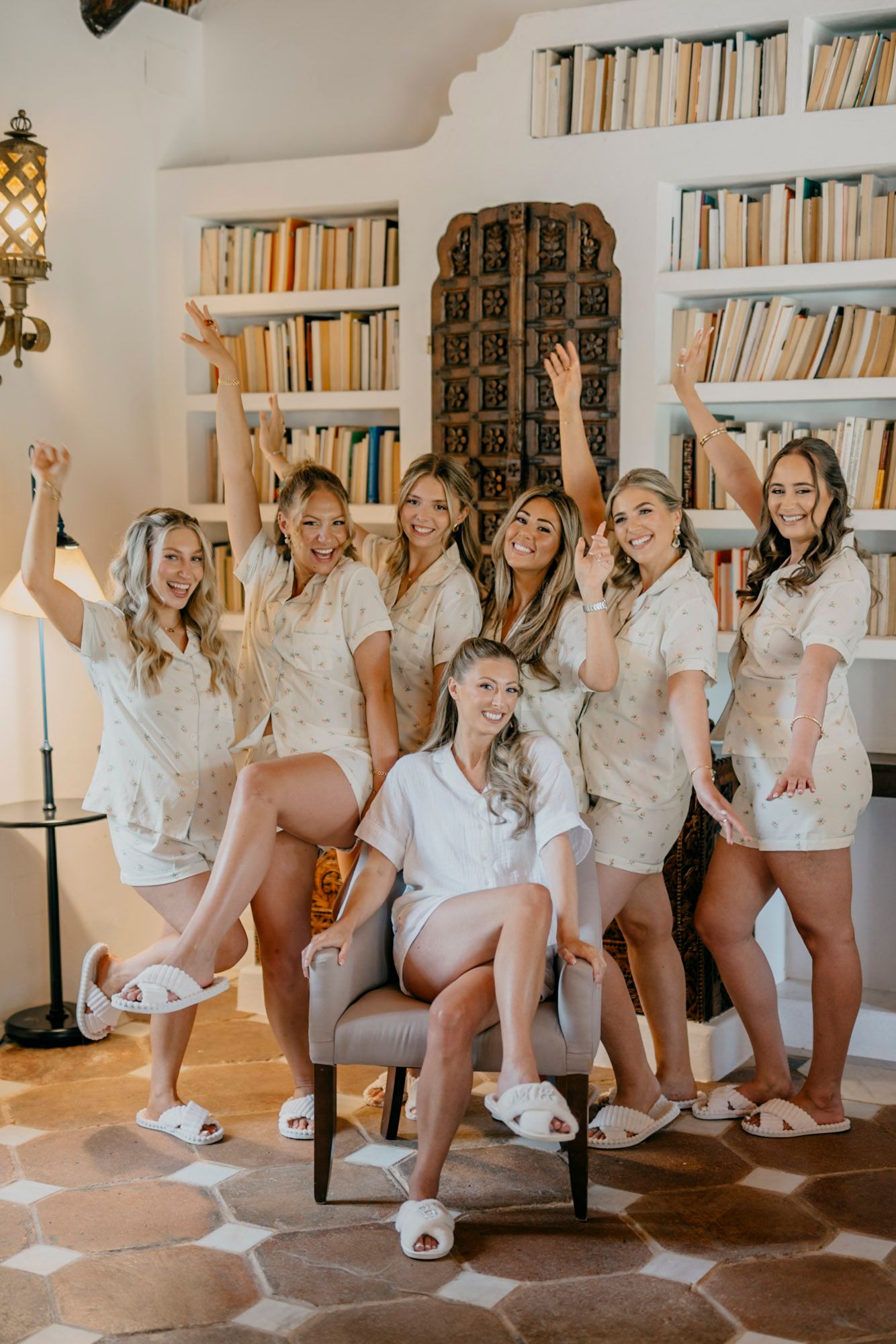 A group in matching white pajama sets poses together in front of built-in bookshelves during a celebratory gathering.