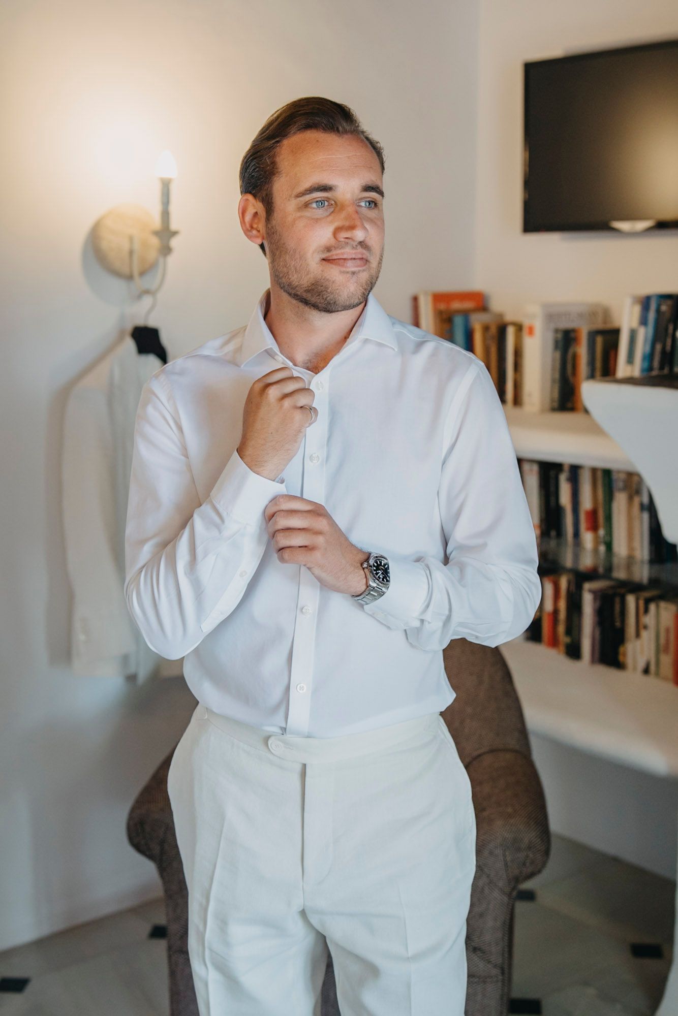 Person in all white formal attire adjusting their collar in a modern room with bookshelves.