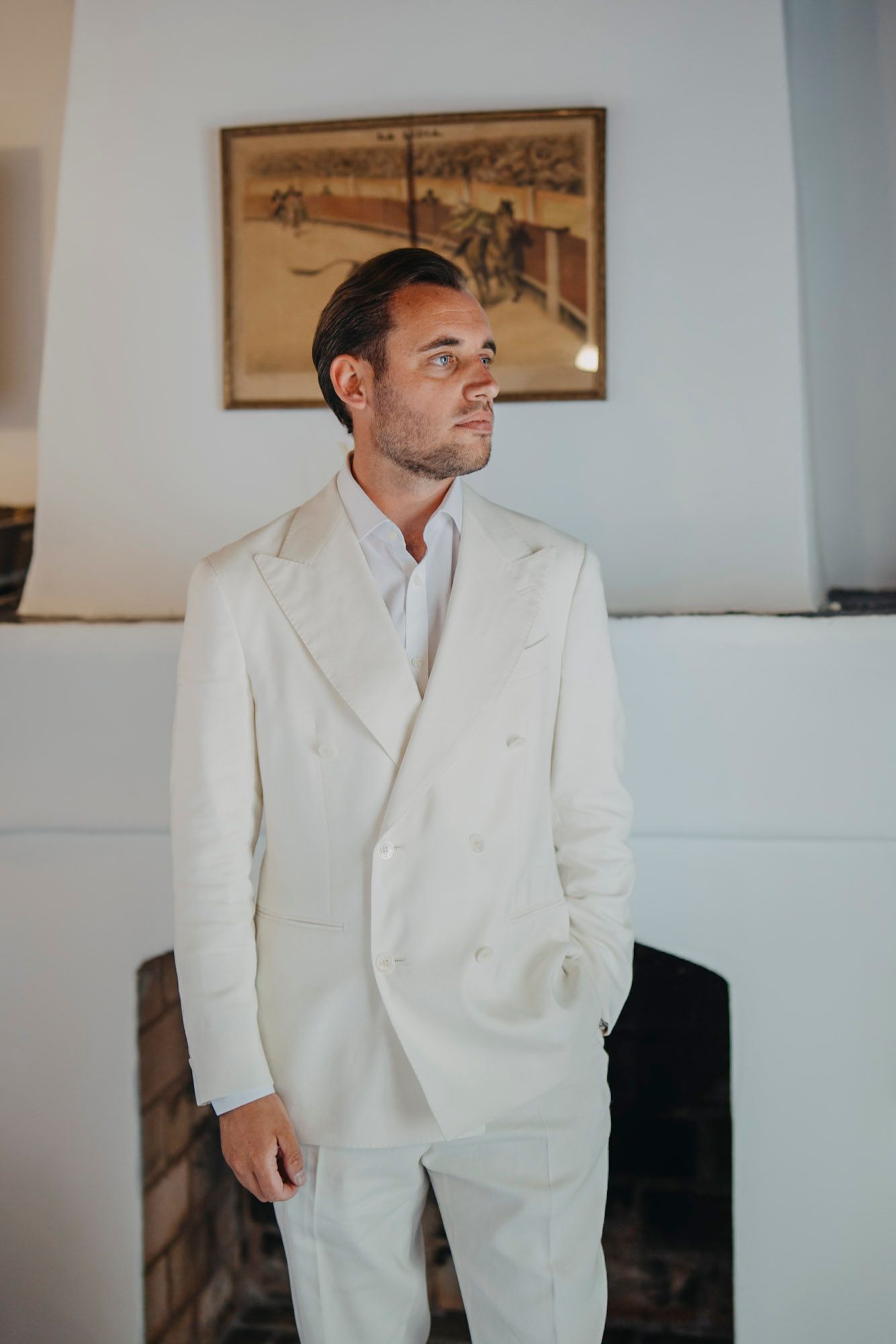 Groom in crisp white suit poses thoughtfully in elegant indoor setting.