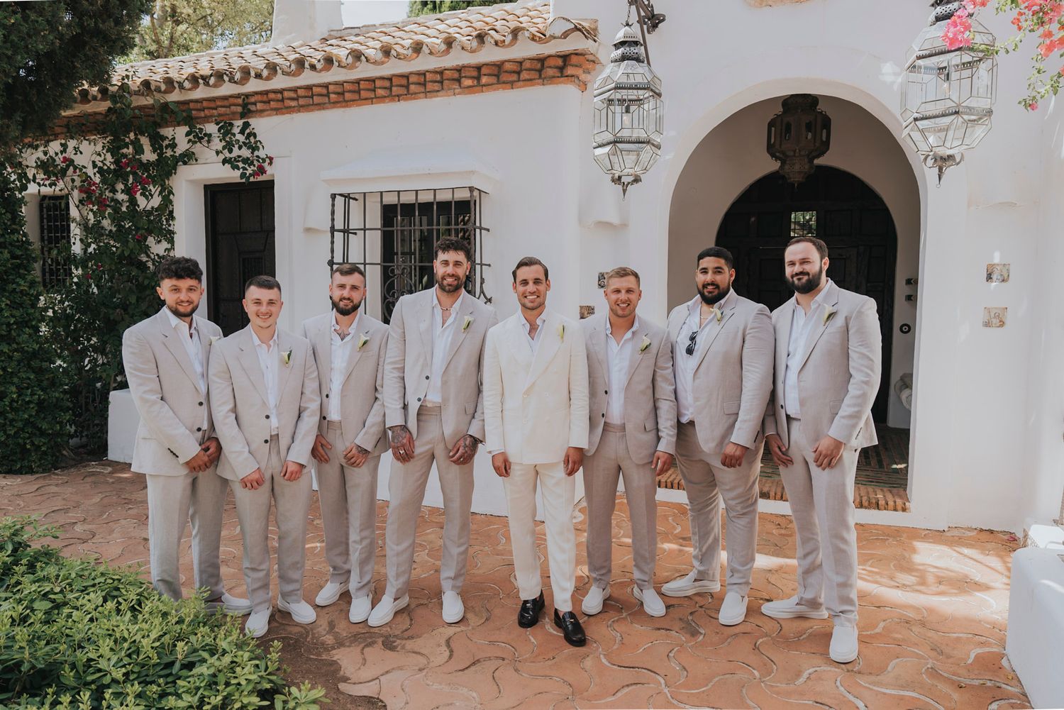 Groomsmen in light beige suits standing together outside Spanish-style building entrance.