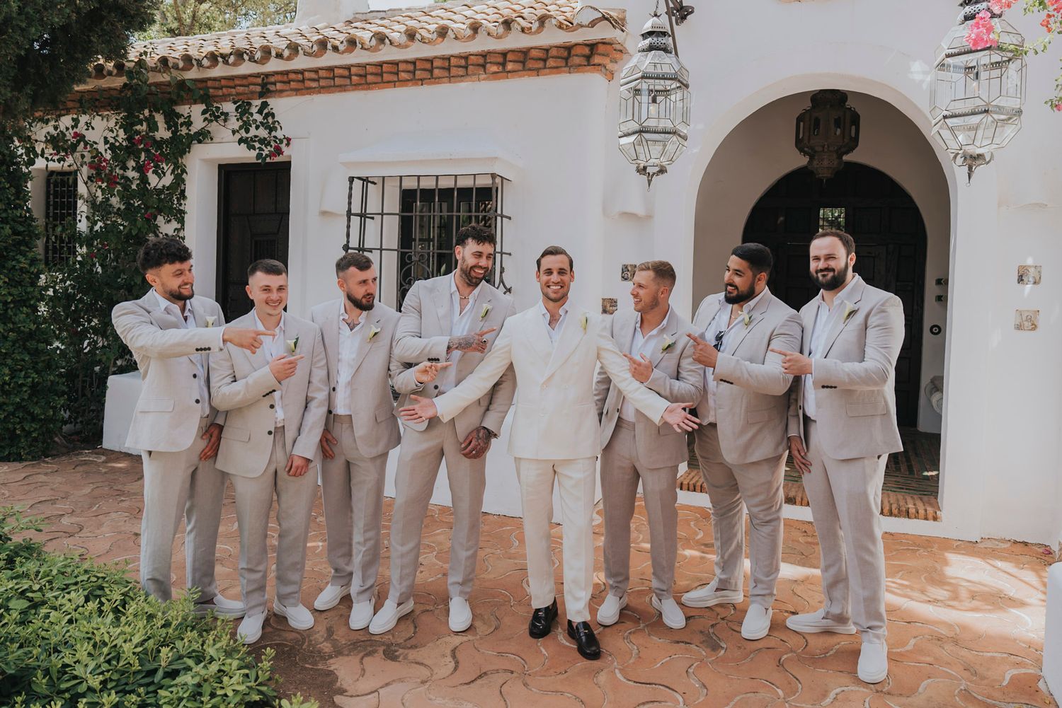 Groomsmen in light beige suits pose together outside a Mediterranean-style building.
