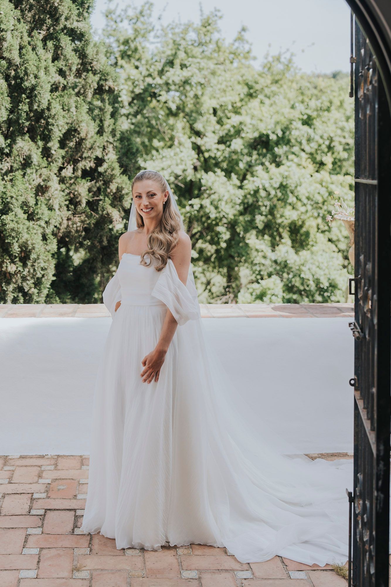 Bride in flowing white gown posing against stone wall in garden setting.
