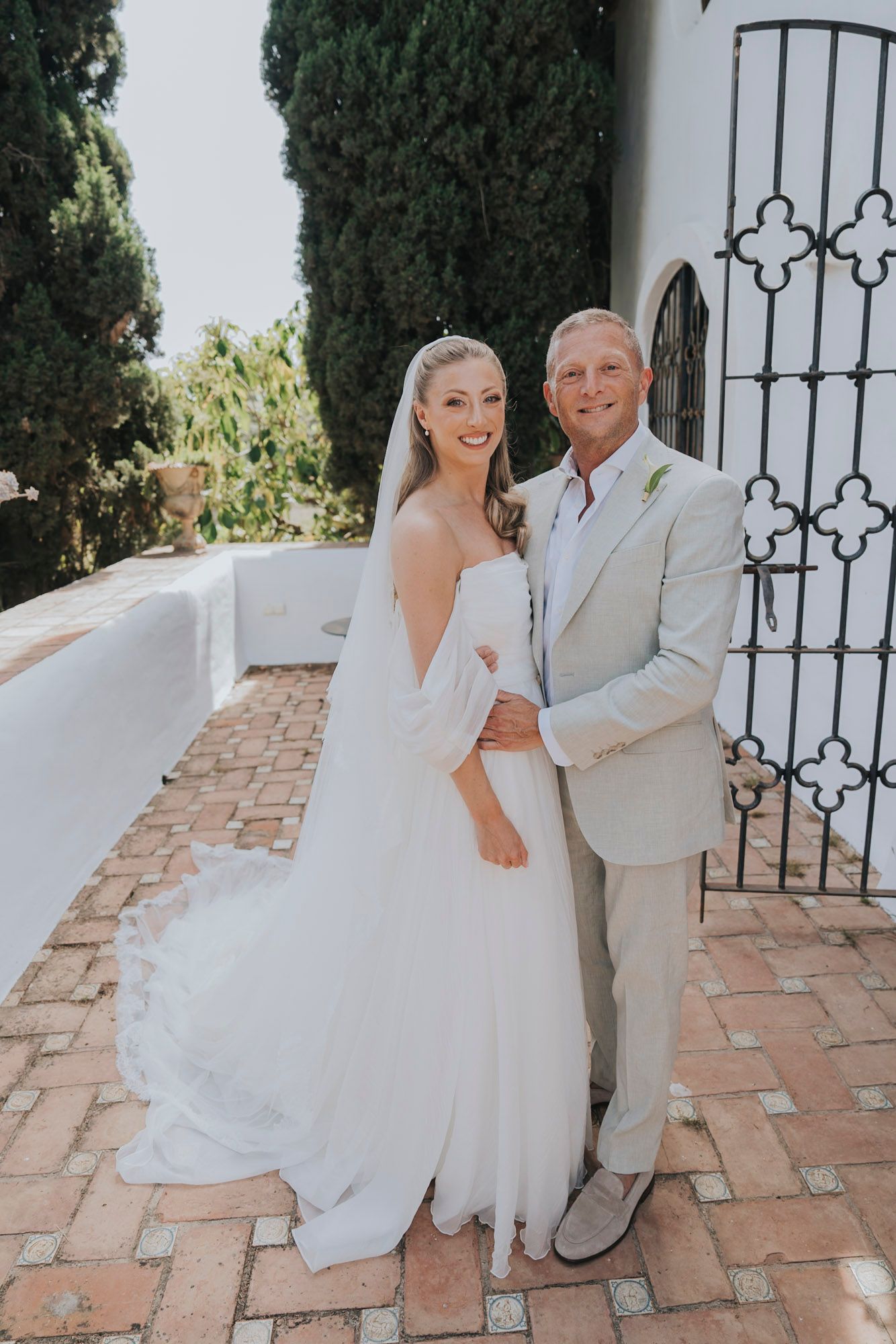 Couple in wedding attire posing playfully by white stucco wall and wrought iron gate.