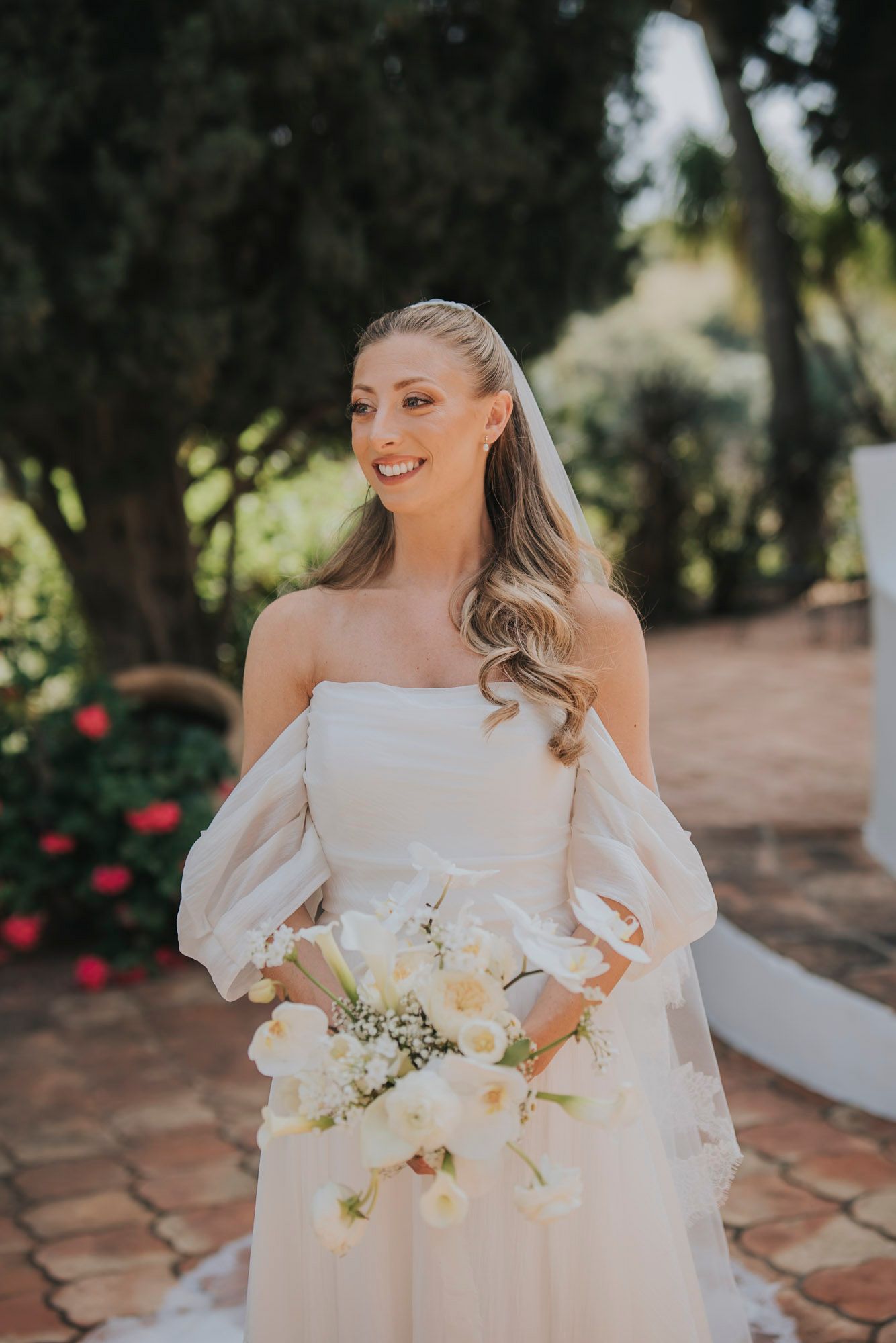 Elegant white wedding dress with puff sleeves and white floral bouquet on terracotta tiles.