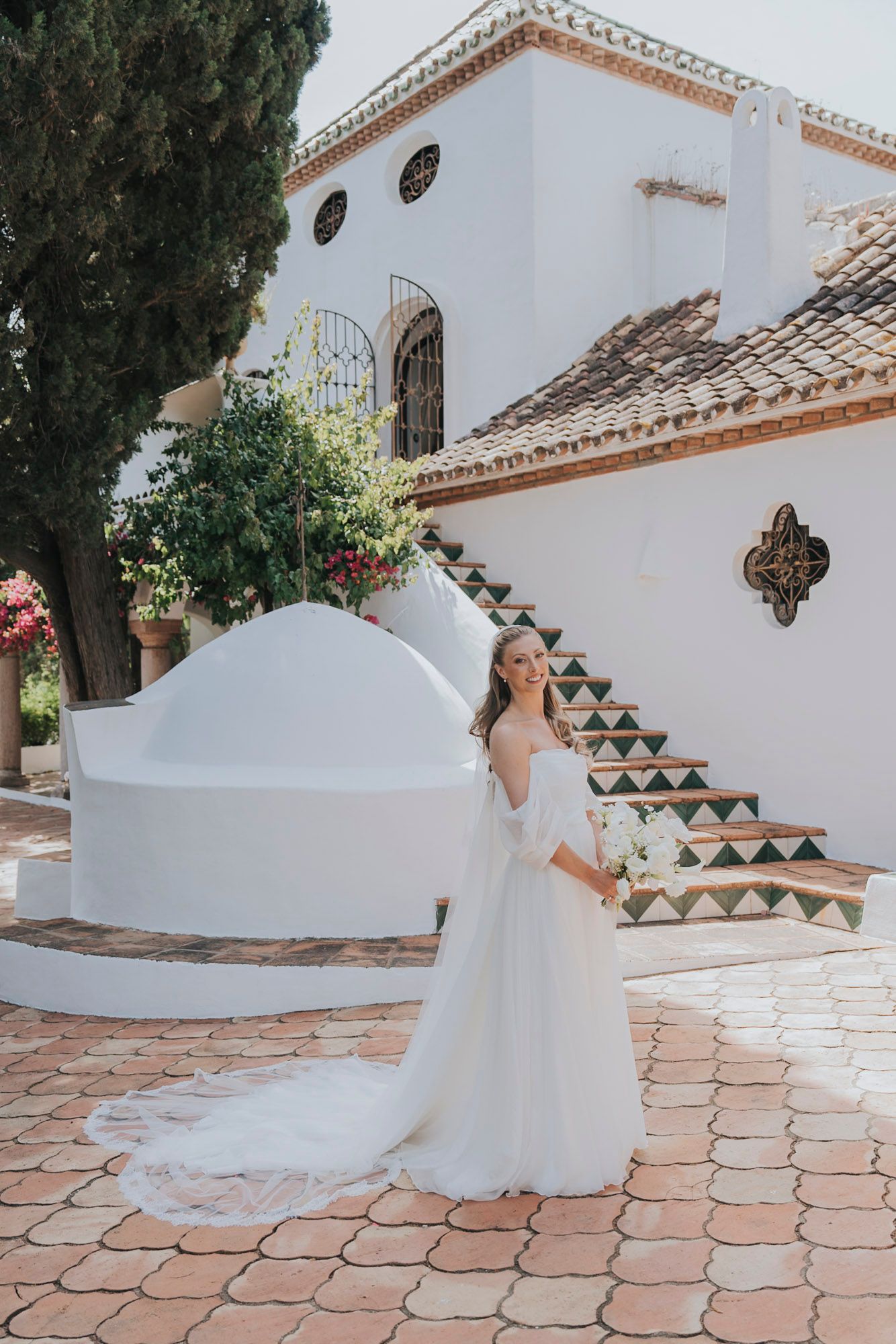 Bride in flowing white dress poses on terracotta tiles at Mediterranean venue.
