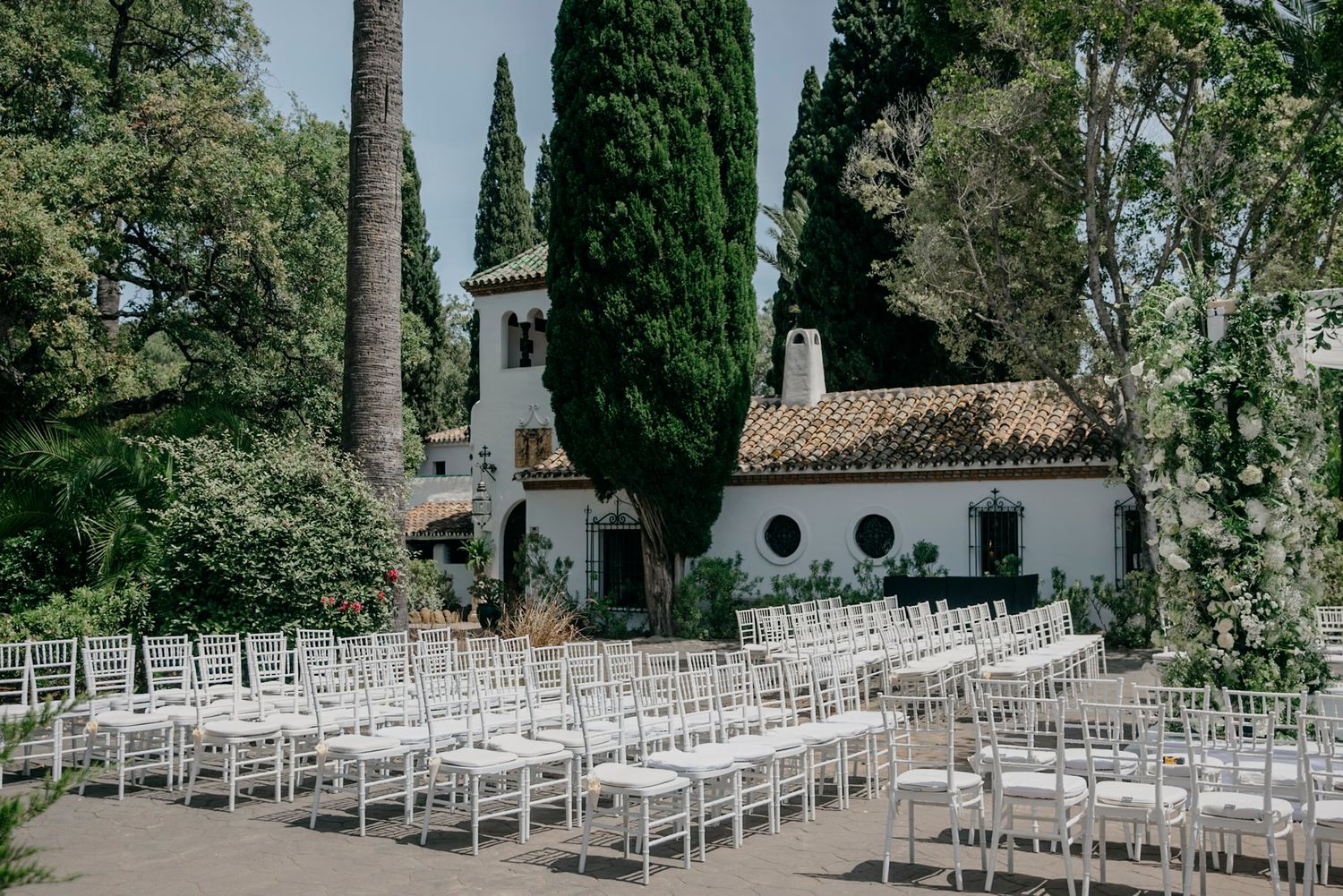 White ceremony chairs arranged in rows on a garden lawn with cypress trees and white architecture.