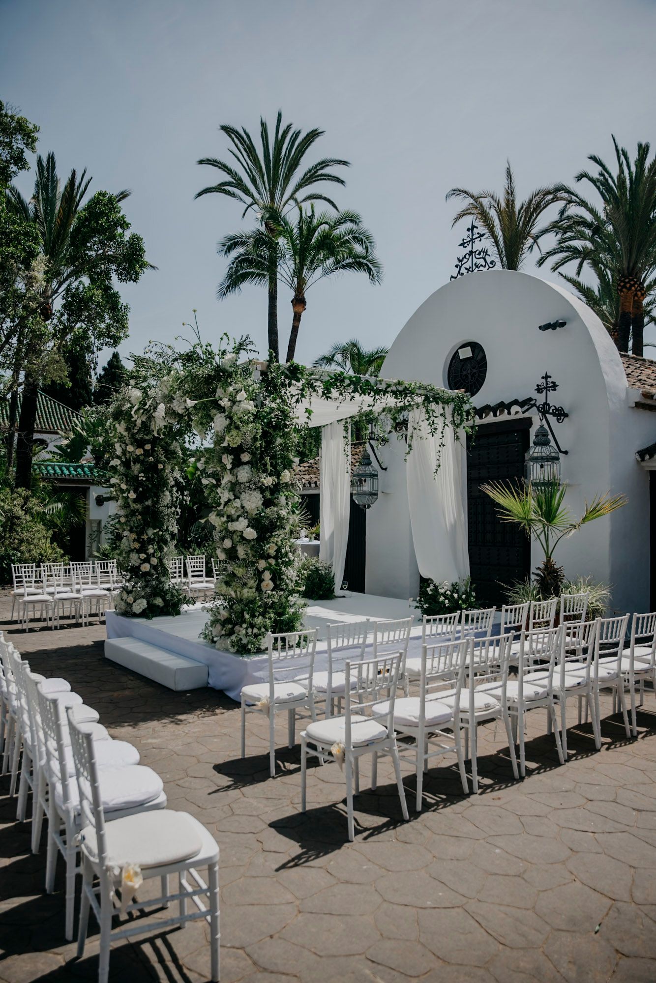 Outdoor wedding ceremony setup with white chairs and domed architecture against palm trees.