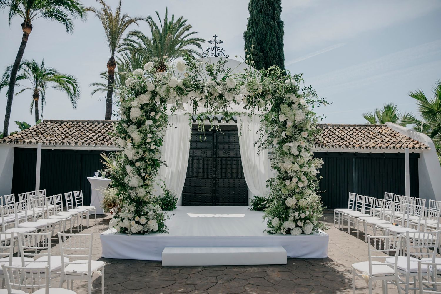 White chairs arranged in rows for an outdoor wedding ceremony with palm trees and greenery.