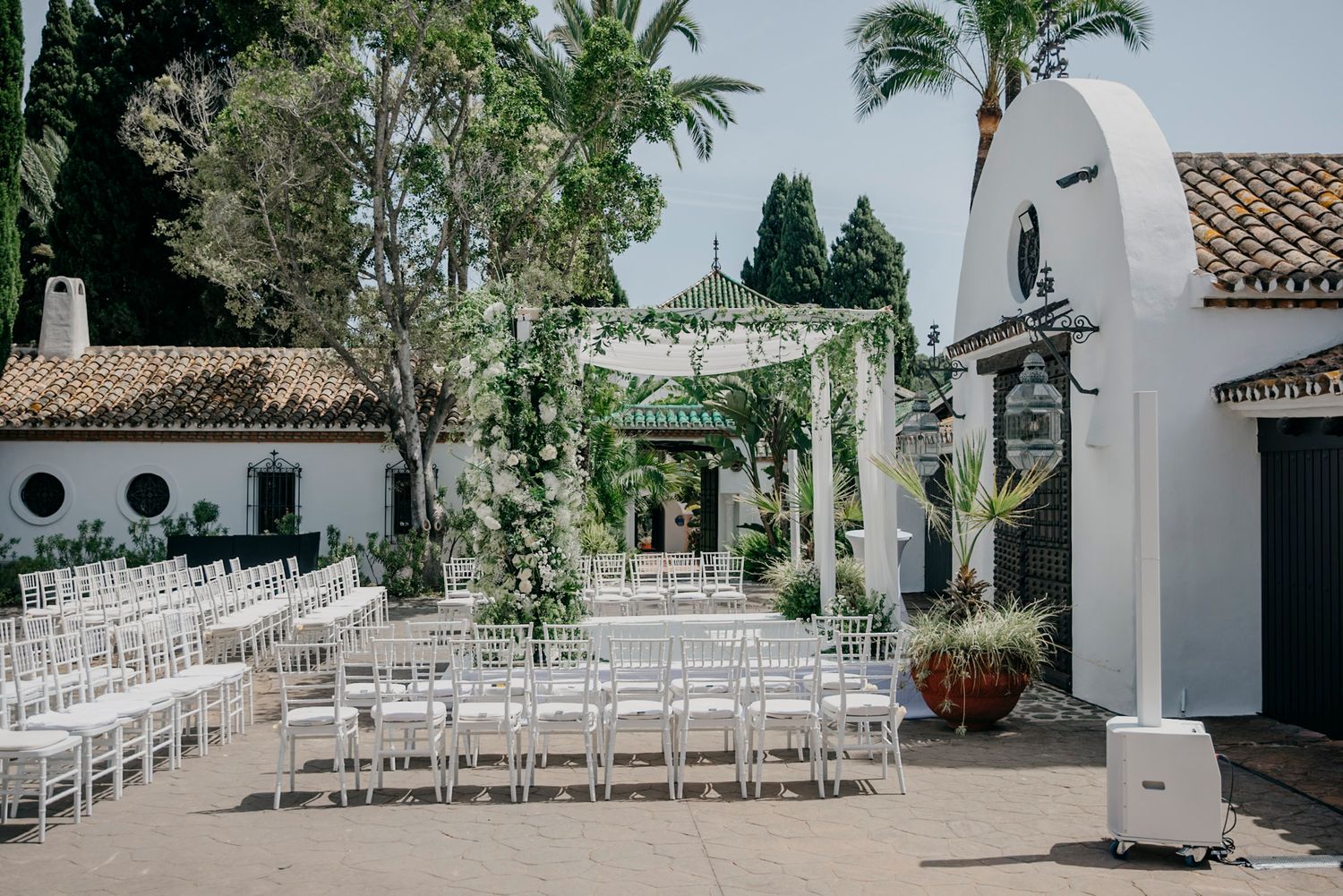 White chairs set up for an outdoor wedding ceremony with palm trees and Spanish architecture.