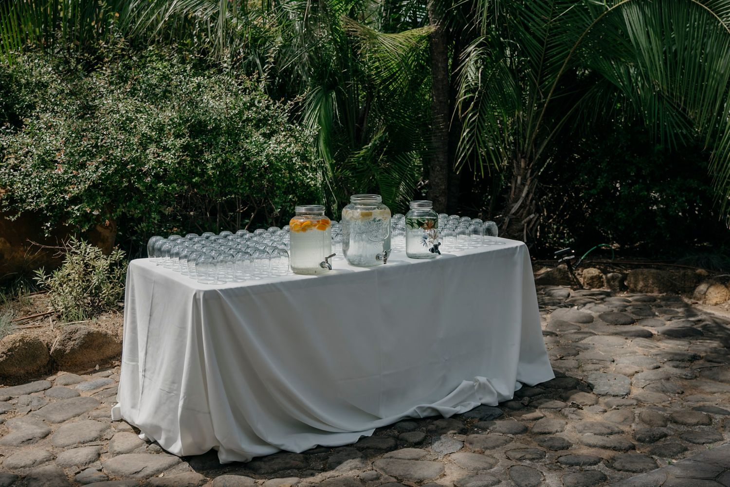 White tablecloths cover tables set up outdoors with palm trees and natural greenery in the background.