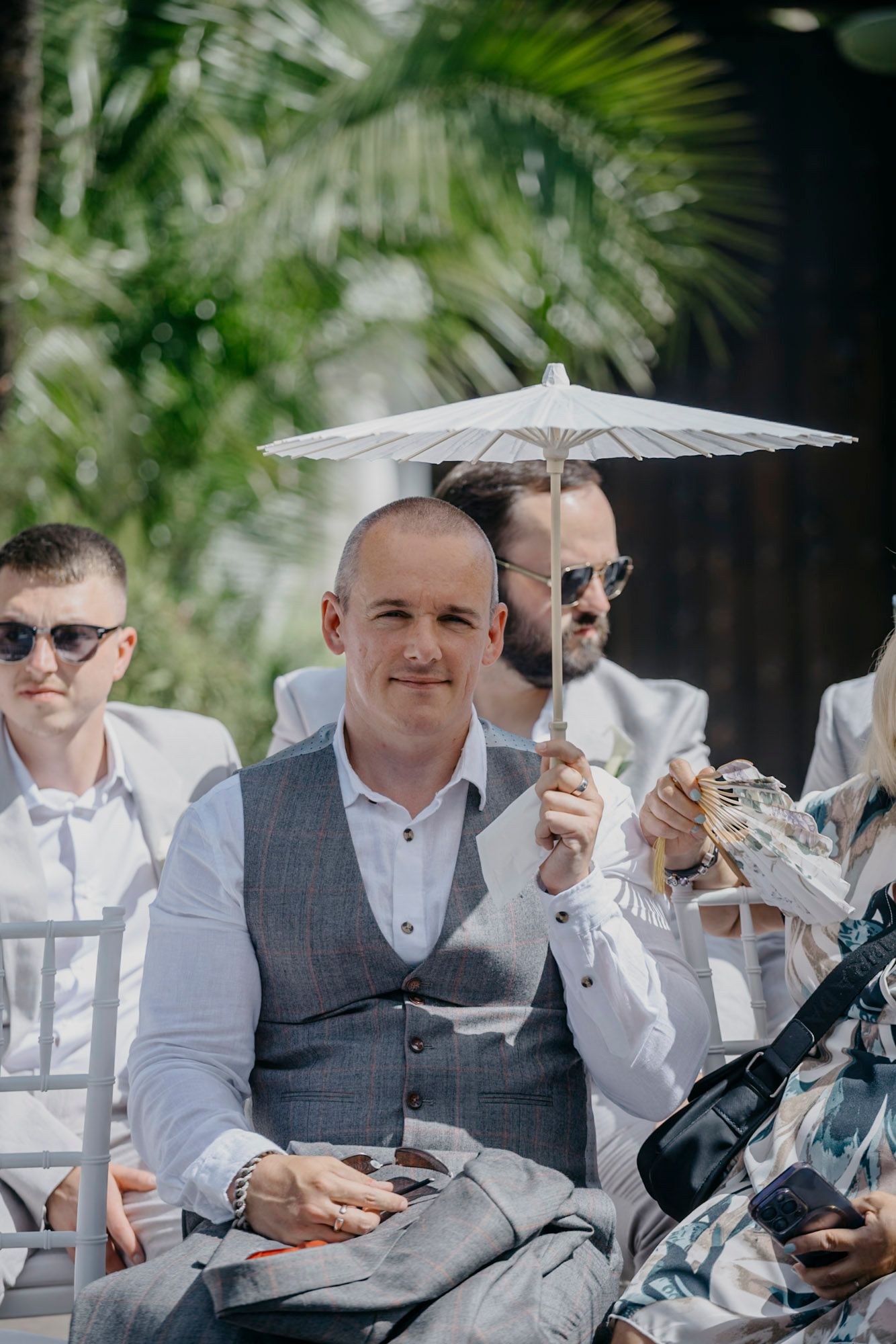 Wedding guests holding parasols while seated in formal attire at an outdoor ceremony.