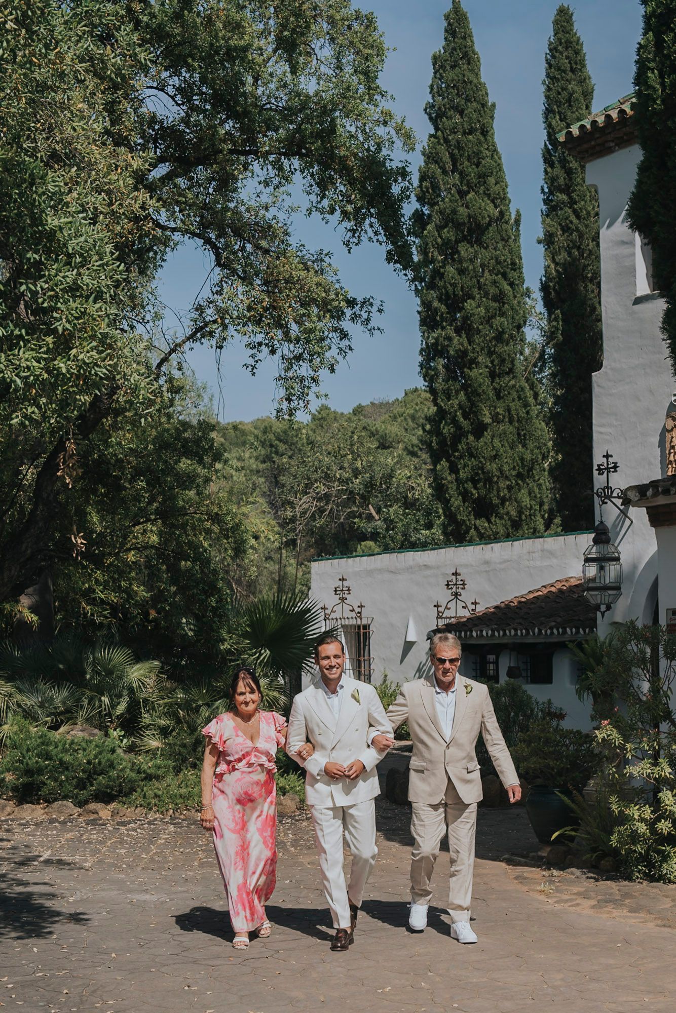 Wedding party walking along a path with cypress trees and white buildings in the background.