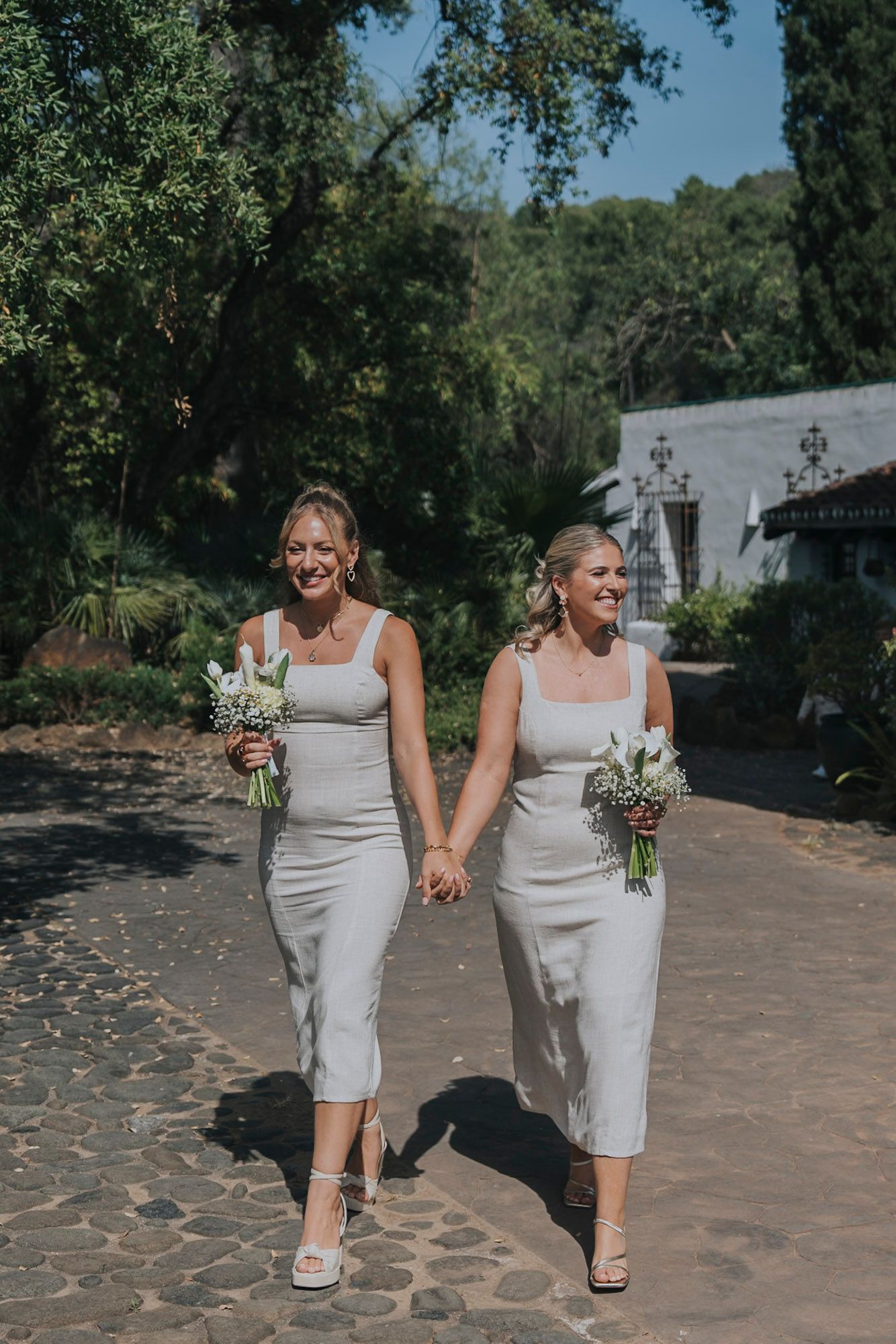 Two people in matching light gray dresses walk together holding bouquets outdoors.