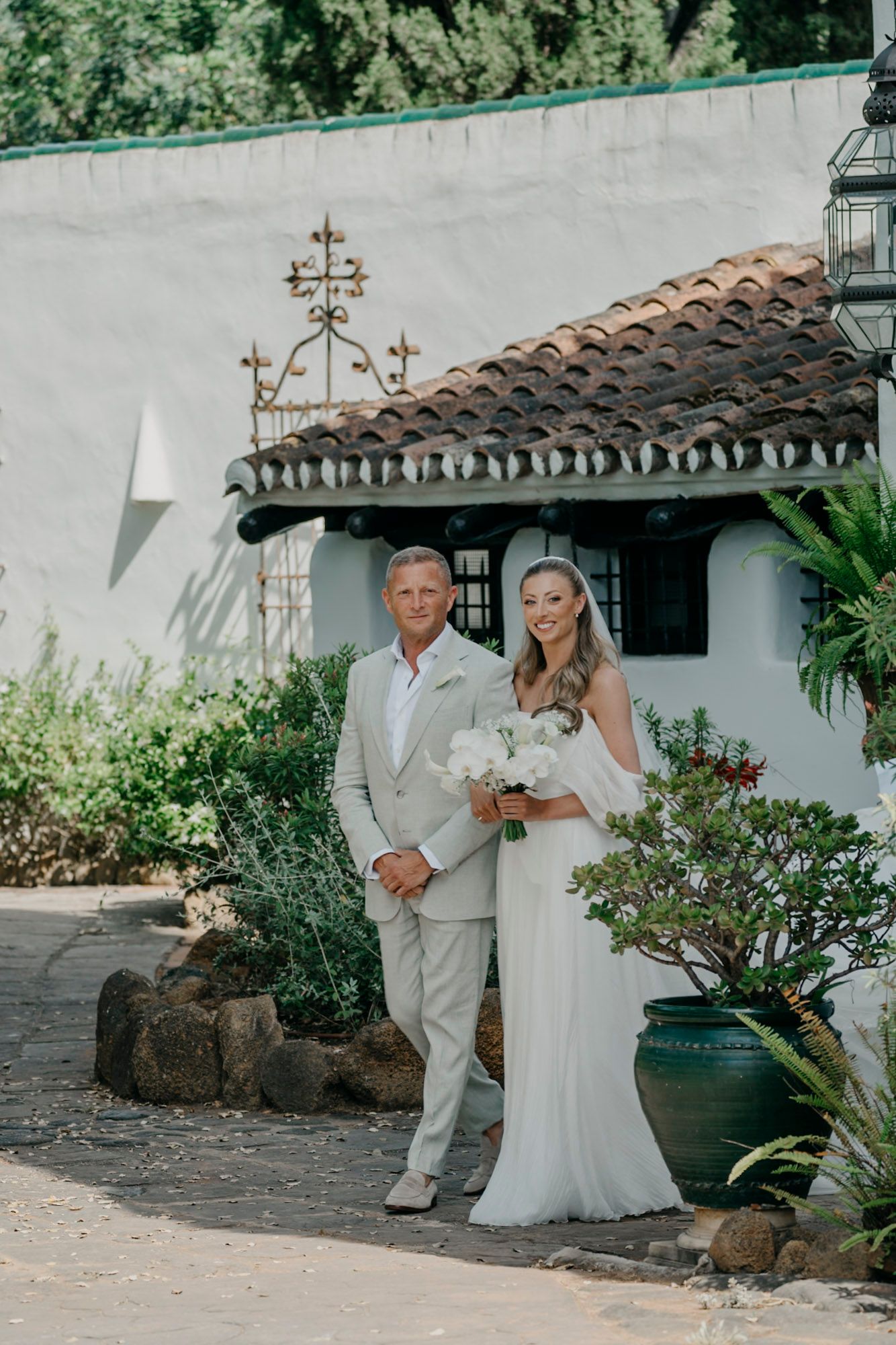 Elegant wedding couple poses against white Spanish colonial architecture with terracotta roof tiles.