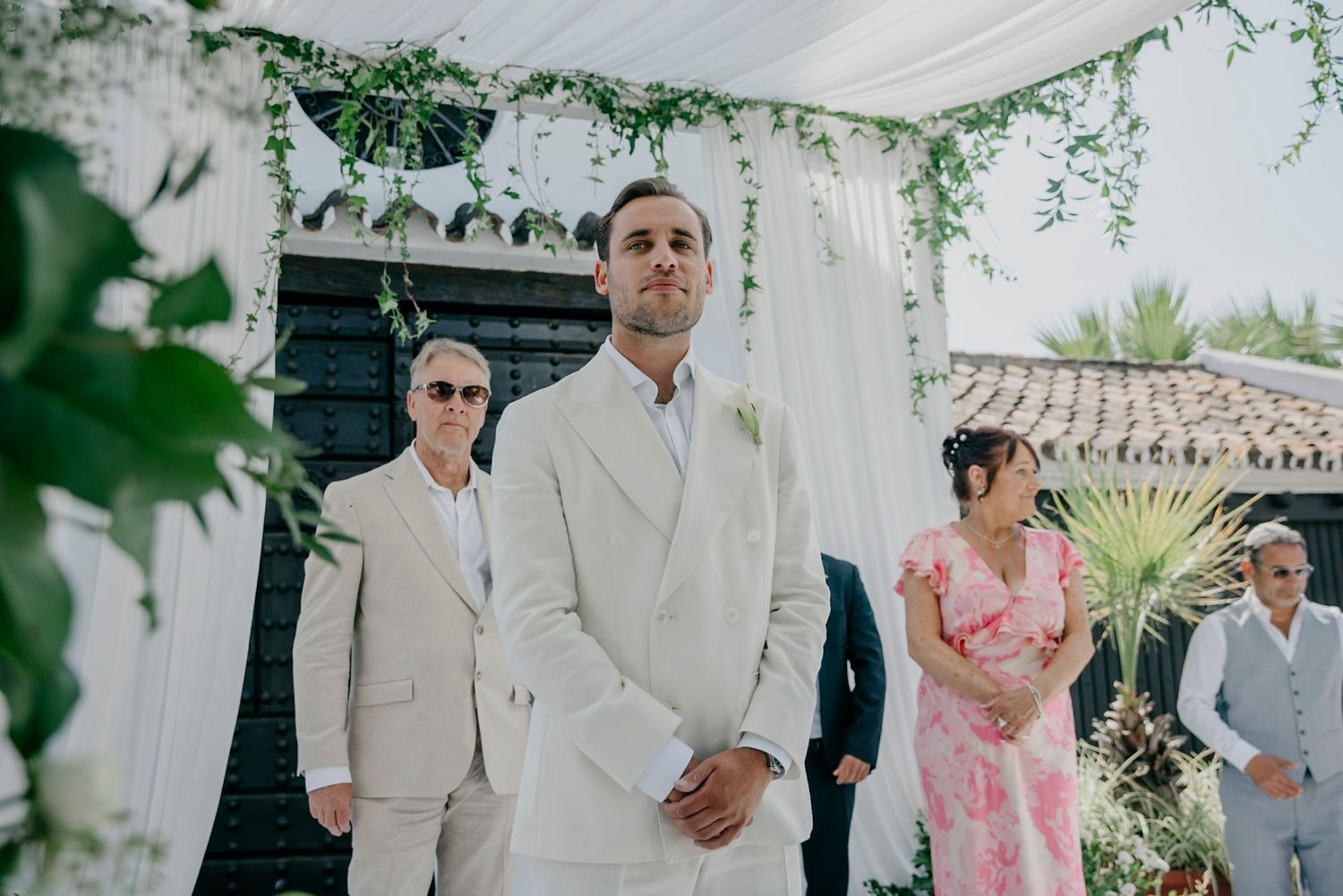 Wedding ceremony guests gather under greenery and floral decorations at an outdoor venue.