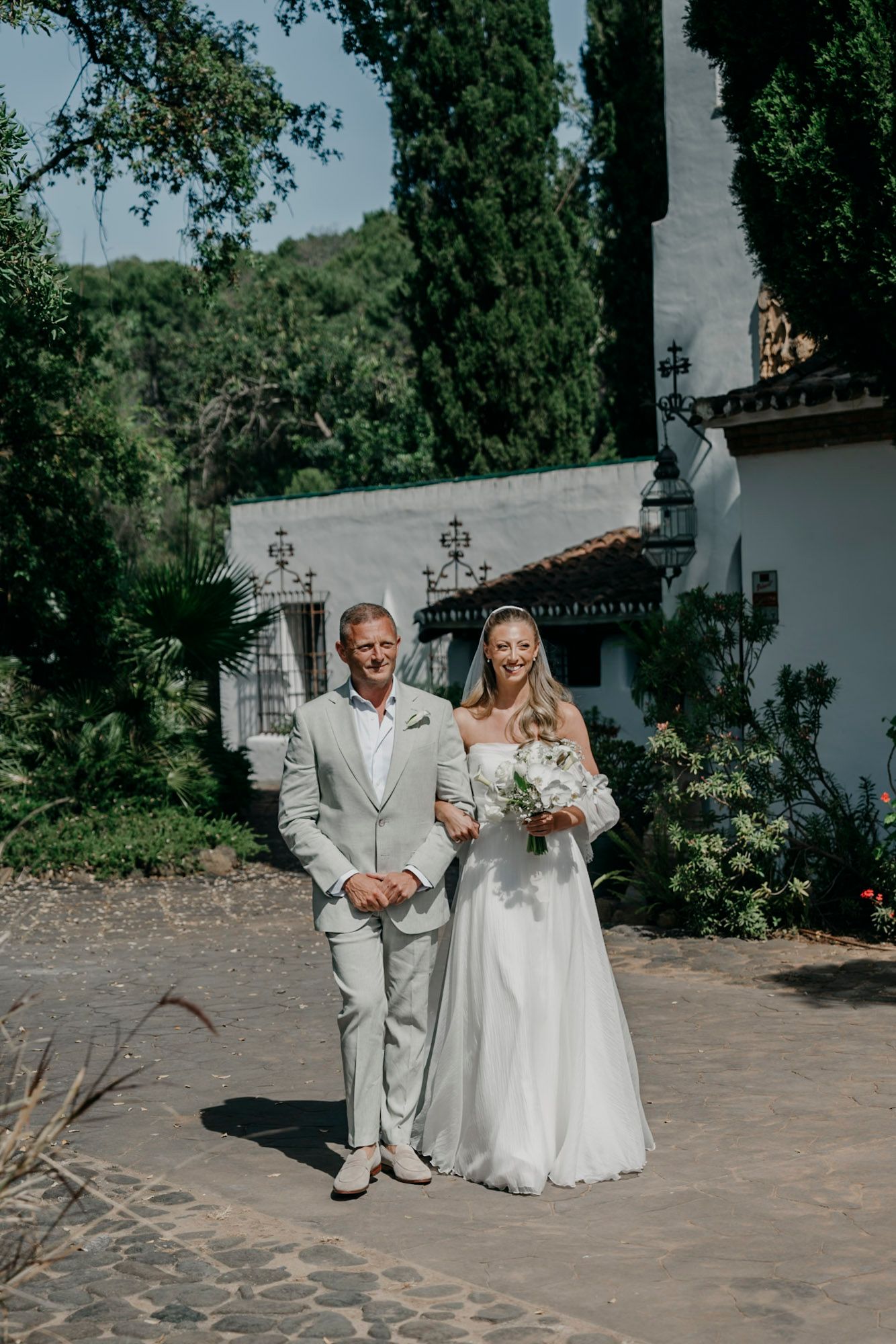 Wedding processional on stone pathway with white stucco walls and garden landscaping.